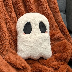 May include: A white plush ghost pillow with black eyes and pink cheeks, sitting on a brown fuzzy blanket.