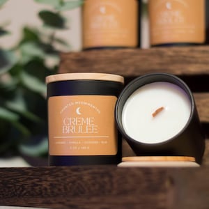 May include: A black candle jar with a wooden lid and a label that reads "Haunted Moments Creme Brulee Caramel, Vanilla, Custard, Rum 8 oz / 240 g". The candle is sitting on a wooden surface.