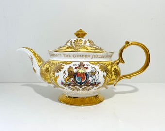 May include: A white teapot with gold trim and a yellow lid. The teapot features a royal coat of arms and the text "Celebrate The Golden Jubilee 1952-2002".