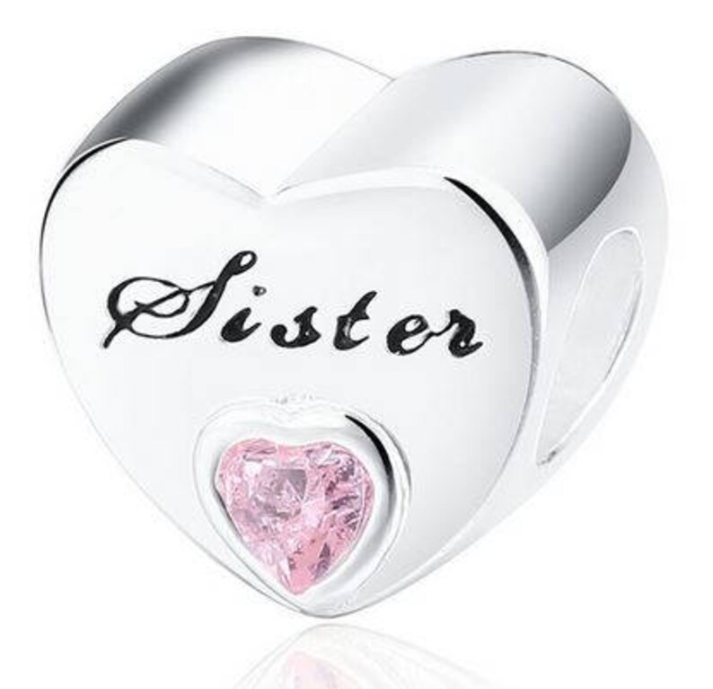 May include: A silver charm in the shape of a heart. The word "Sister" is written in black script across the front. A smaller heart-shaped pink gem is set in the center of the charm.