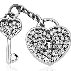 Puede incluir: Un colgante plateado con forma de corazón con cerradura y llave. La cerradura y la llave están conectadas por una cadena. Tanto la cerradura como la llave están incrustadas con pequeñas piedras preciosas transparentes. La llave tiene un mango en forma de corazón.