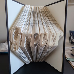 May include: A book with the word "STOLEN" folded into the pages. The book is open and standing upright on a black surface.