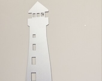 May include: A silver metal silhouette of a lighthouse with a small house next to it. The lighthouse has a tall tower with four windows and a small roof. The house has one window and a small roof. Both are on a base that looks like a rocky shore.