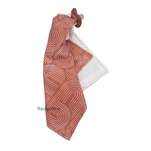 May include: A baby pacifier attached to a rust-colored fabric with a white, curved line pattern. The fabric is folded over a white square, creating a soft, textured baby item. The pacifier is a light brown color.