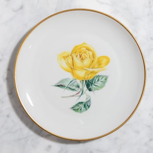 May include: A white ceramic plate with a gold rim, decorated with a yellow rose and green leaves. The plate is on a marble surface. The design is classic and elegant. The plate is suitable for serving food.