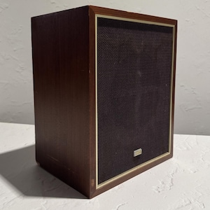 May include: A rectangular speaker cabinet with a dark brown wood veneer finish. The front features a dark brown fabric grille framed by a thin light-colored border, with a small rectangular metallic logo badge centered at the bottom.