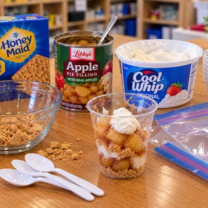 Pode incluir: Uma caixa de bolachas Honey Maid, uma lata de recheio de tarte de maçã Libby's, um recipiente de Cool Whip, um copo de plástico transparente com camadas de migalhas, recheio de maçã e natas, uma taça de vidro com migalhas e colheres de plástico.