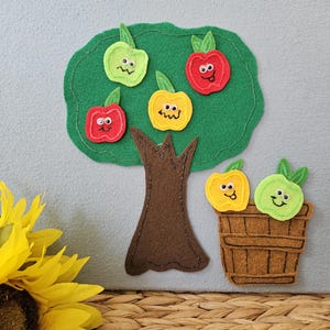 May include: A felt craft set featuring a brown tree with a green canopy and four colorful apples with googly eyes and stitched faces. A brown basket sits beside the tree containing two additional apples with stitched faces and googly eyes.