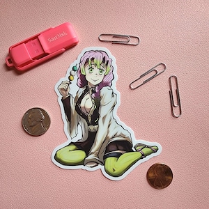 May include: A sticker featuring a cartoon anime character with pink and green hair, wearing a white and black outfit, holding a skewer of green and white dango. The character is sitting with their legs crossed and has a light green background.
