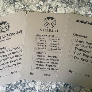 May include: Three beige file folders with text and logos from the Avengers Initiative, S.H.I.E.L.D., and Stark Industries. Each folder has a list of contents and a place for a date and signature. The folders are for incident reports and other records.