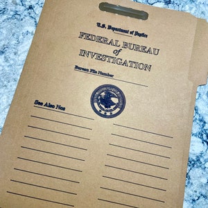 May include: A tan file folder with the text "U.S. Department of Justice FEDERAL BUREAU of INVESTIGATION" in black. The folder has a handle cutout and a seal. Lines are printed below the text "See Also Nos".