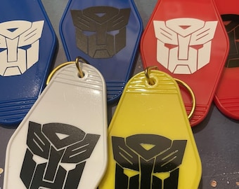 May include: A collection of colorful keychains in blue, red, white, and yellow. Each keychain features a black and white graphic design. The keychains have a retro motel style shape and are attached to gold-colored key rings.