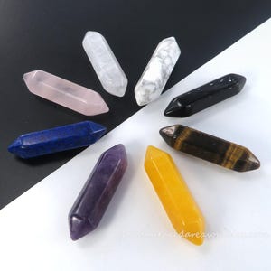 May include: An assortment of double-terminated crystal wands in various colors, including rose quartz, amethyst, and tiger's eye. The crystals are arranged in a circular pattern against a black and white background. The Etsy shop name is visible.