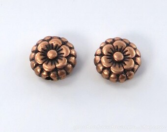 May include: Two bronze-colored, flower-shaped beads. Each bead has a central flower design surrounded by multiple petals. The beads are round and have a slightly textured surface. The background is plain white.