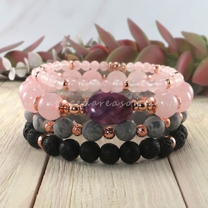 May include: Stack of four beaded bracelets. The top two bracelets feature pink beads and rose gold accents. The third bracelet has gray beads and a large purple stone. The bottom bracelet has black lava beads.