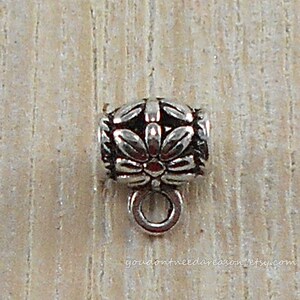 May include: Silver-toned metal bead with a floral design and a small loop at the bottom. The bead is on a wooden surface.