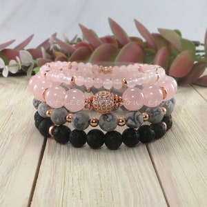 May include: Four stacked bracelets with rose quartz, grey and black lava beads. The top bracelet has a rose gold charm with clear rhinestones.