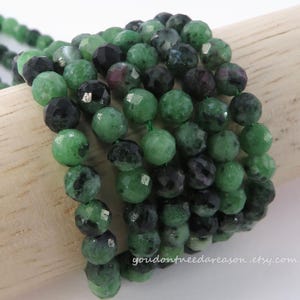 May include: Close-up of a beaded bracelet featuring faceted green and black beads. The beads are strung together on a green cord. The Etsy shop name "youdontneedareason.etsy.com" is visible.