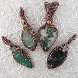 May include: Four handcrafted gemstone pendants with copper wire wrapping. The pendants feature various green and black stones, including a clear quartz crystal. The wirework is intricate, with decorative swirls and bead accents. The pendants are displayed on a textured white surface.