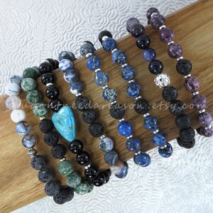 May include: Six lava stone bracelets with different colored gemstone beads. The bracelets feature a variety of colors, including blue, green, purple, and white. One bracelet has a turquoise heart charm.