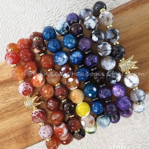 May include: A collection of beaded bracelets in a rainbow of colors, including red, orange, yellow, blue, and purple. Each bracelet features large, round beads and gold-colored accents. The bracelets are adorned with small, gold-colored crown charms. The text "you don't need a reason" is visible.