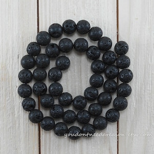 May include: A necklace of black lava rock beads, each with a porous texture, arranged in a circular shape. The beads are set against a light wood grain background. The Etsy shop name "youdontneedareason.etsy.com" is visible.