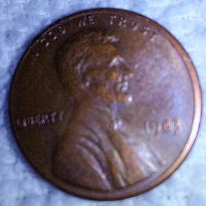 May include: A copper-coloured United States penny from 1983, featuring a profile of a man and the inscriptions "IN GOD WE TRUST" and "LIBERTY". The coin is circular and shows signs of wear.