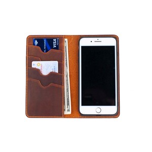 Puede incluir: Una cartera de cuero marr&oacute;n con una funda para tel&eacute;fono y m&uacute;ltiples ranuras para tarjetas. La cartera est&aacute; abierta y muestra una tarjeta de cr&eacute;dito, una tarjeta de d&eacute;bito y un billete de d&oacute;lar.