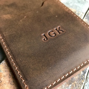 May include: A brown leather wallet with a stitched edge and the initials "JGK" embossed on the front.