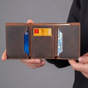 May include: A brown leather wallet with multiple card slots and a billfold. The wallet is open and shows a Wells Fargo Visa card, a blue card with a white logo, and a New York State driver's license.