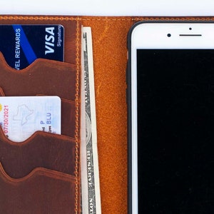 Puede incluir: Una cartera de cuero marr&oacute;n con m&uacute;ltiples ranuras para tarjetas, un bolsillo para efectivo y un soporte para tel&eacute;fono. La cartera tiene un borde cosido y un tel&eacute;fono blanco con una pantalla negra est&aacute; insertado en el soporte para tel&eacute;fono.