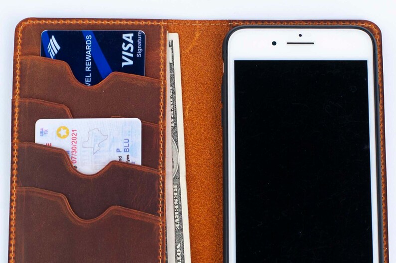 Puede incluir: Una cartera de cuero marr&oacute;n con m&uacute;ltiples ranuras para tarjetas, un bolsillo para efectivo y un soporte para tel&eacute;fono. La cartera tiene un borde cosido y un tel&eacute;fono blanco con una pantalla negra est&aacute; insertado en el soporte para tel&eacute;fono.