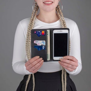 Puede incluir: Una funda de tel&eacute;fono de cuero negro con m&uacute;ltiples ranuras para tarjetas y un bolsillo para efectivo. La funda est&aacute; abierta y se puede ver un tel&eacute;fono inteligente en el interior. La funda est&aacute; en manos de una persona.