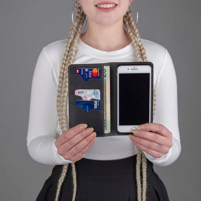 Puede incluir: Una funda de tel&eacute;fono de cuero negro con m&uacute;ltiples ranuras para tarjetas y un bolsillo para efectivo. La funda est&aacute; abierta y se puede ver un tel&eacute;fono inteligente en el interior. La funda est&aacute; en manos de una persona.