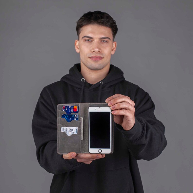 Puede incluir: Una funda de tel&eacute;fono de cuero gris con una ranura para tarjetas de cr&eacute;dito y efectivo. La funda est&aacute; abierta y se puede ver un tel&eacute;fono inteligente negro en el interior.