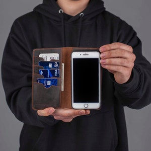 Puede incluir: Una funda de tel&eacute;fono de cuero marr&oacute;n con una ranura para tarjetas de cr&eacute;dito y un soporte de tel&eacute;fono incorporado. La funda est&aacute; abierta y muestra un tel&eacute;fono inteligente negro y varias tarjetas de cr&eacute;dito.