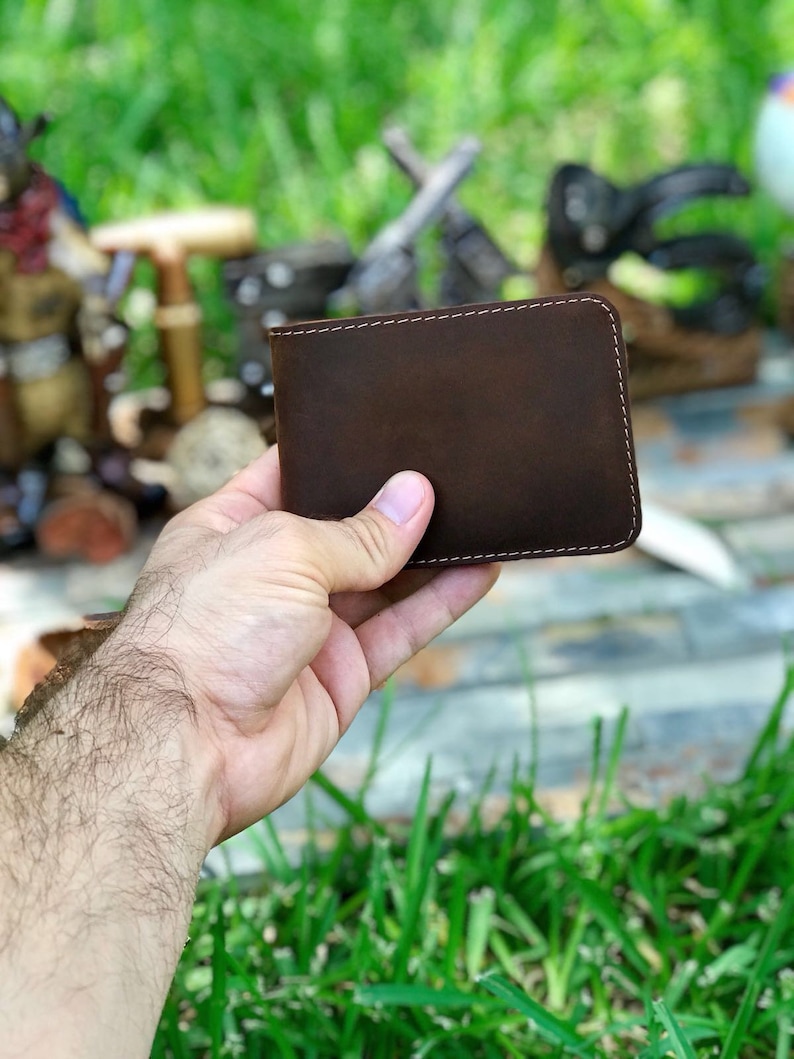May include: A brown leather wallet with white stitching. The wallet is closed and being held in a hand.