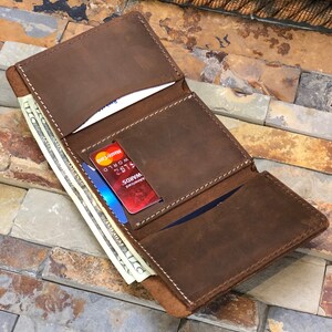 May include: A brown leather trifold wallet with a stitched edge, open to reveal three card slots and a cash pocket. The wallet is filled with cash and credit cards, including a red Visa card and a blue Mastercard.