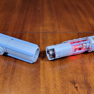 May include: A disassembled lightsaber prop with a light blue outer shell and a red light inside. The lightsaber is made of plastic and has a metal screw on the end.