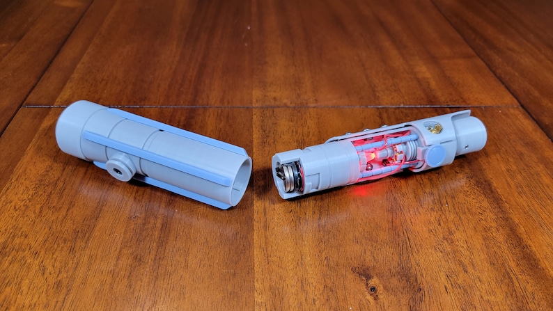 May include: A disassembled lightsaber prop with a light blue outer shell and a red light inside. The lightsaber is made of plastic and has a metal screw on the end.