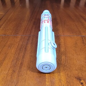 May include: A light blue plastic toy lightsaber with red details. The lightsaber is lying on a wooden surface.