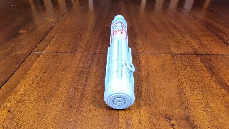 May include: A light blue plastic toy lightsaber with red details. The lightsaber is lying on a wooden surface.