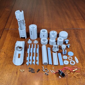 May include: A set of grey 3D printed parts for a lightsaber, including the hilt, blade, and various other components. The parts are laid out on a wooden surface, along with small screws, wires, and other hardware.