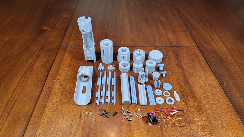 May include: A set of grey 3D printed parts for a lightsaber, including the hilt, blade, and various other components. The parts are laid out on a wooden surface, along with small screws, wires, and other hardware.