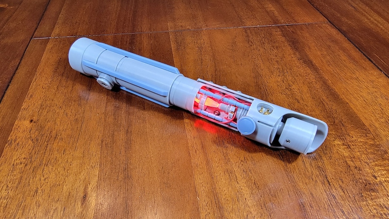 May include: A disassembled lightsaber with a blue and grey plastic exterior. The interior is visible, revealing red wiring and a red LED light.