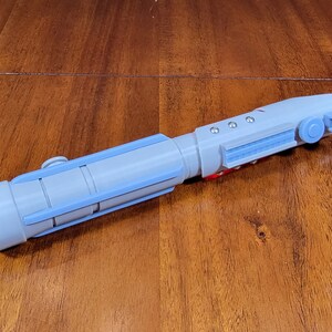 May include: A light blue lightsaber prop with a red button and a black button on the handle. The lightsaber is made of plastic and has a textured surface.