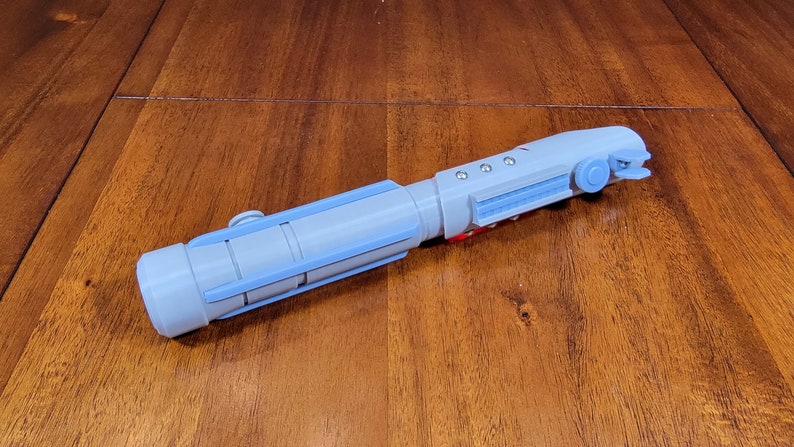 May include: A light blue lightsaber prop with a red button and a black button on the handle. The lightsaber is made of plastic and has a textured surface.