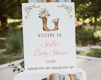 May include: A white sign with gold accents and a floral design featuring two brown cowboy boots. The sign reads "Welcome to Stella's Baby Shower 7.15.2021".