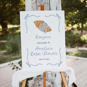 May include: A white sign with blue ribbon accents and a watercolor illustration of a croissant. The sign reads "Bonjour Welcome to Amelia's Bebe Shower May 10, 2025".