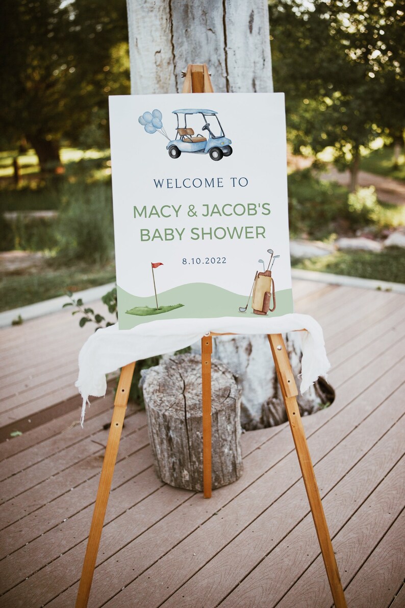 May include: A white sign with a blue golf cart and golf clubs, announcing a baby shower for Macy & Jacob on 8.10.2022. The sign is on a wooden easel.
